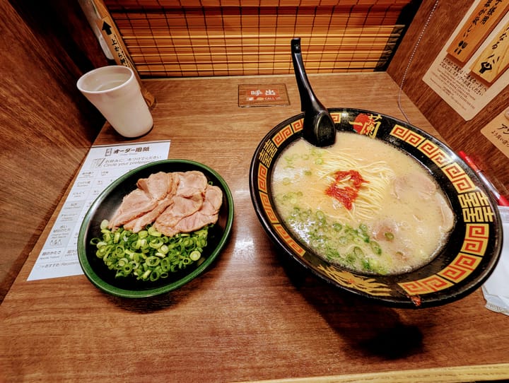 A very small cubby, with a large bowl of ramen on the right, and extra pork and green onions on the left. A screen in the back that opens when being served.