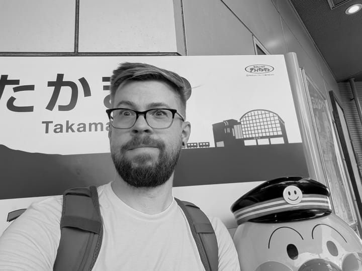 Black and white photo of a male taking a selfie next to a "plastic?" statue of a cartoonish train conductor also sitting on the same bench.